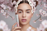 Model holding La Paz Cosmetics Crystal Frosting Lip Gloss in Frosted Blush D19 with sheer pink shimmer lips and cherry blossoms in the background