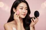 Woman applying Fresco Olive with a pink sponge and compact on a pink background