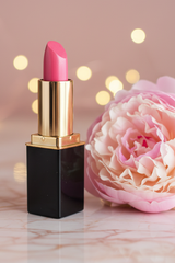 #03 Azalea lipstick with a black square tube and gold collar stands on a marble surface next to a large pink peony flower, with soft golden bokeh lights in the background.