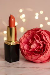 #06 Harvest lipstick with a black and gold accents stands on a light marble surface beside a large pink flower, with warm blurred fairy lights in the background.