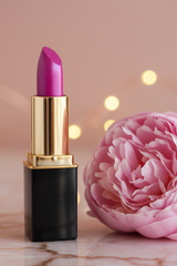 A lipstick tube in L'Paige #21 Electric Fuschia with a gold collar, and black base, styled next to a large pink peony flower on a marble surface with warm bokeh lights in the background