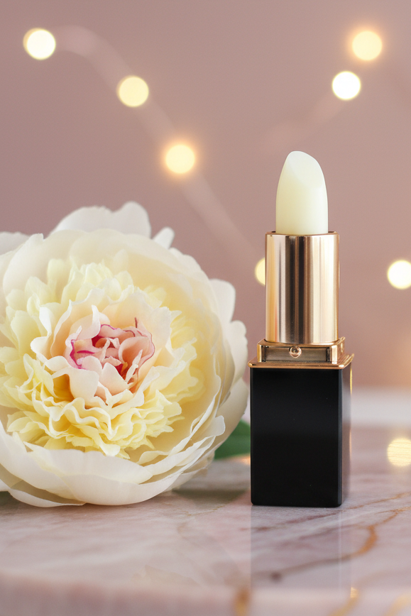An Aloe Treatment Stick in a  lipstick tube with a black square base and shiny gold collar stands beside a large pale cream flower on a marble surface, with warm blurred string lights in the background.