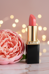 #39 L'Paige lipstick with a gold tube and black base stands on a marble surface next to a large pink flower, with warm bokeh fairy lights in the background.