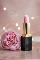 L'Paige #44 Shimmering Pink Ice lipstick with a black and gold tube stands on a pink marble surface next to a pink flower, with soft fairy lights blurred in the background.