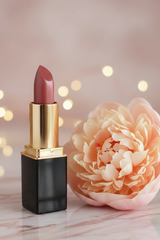 L'Paige Lipstick in #49 Peach Nectar with a black case and gold collar standing beside a large pale peach flower on a marble surface, with soft warm bokeh lights in the background.