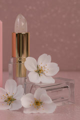 La Paz Cosmetics Blushing Cherry Blossom Crystal Color Changing Lipstick in a clear tube with a gold collar and a translucent white lipstick that changes to you ph shades of pink on a mauve surface beside white blossoms.