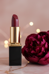 A L'Paige lipstick in color Plum Red with shiny gold collar, and black tube, standing on a marble surface next to a large dark pink peony flower against a soft pink background with warm bokeh lights.