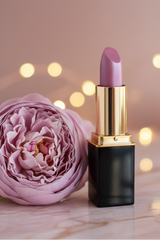 A lipstick tube of #53 Amethyst Mist with a black and gold tube standing beside a large pink flower on a marble surface with warm bokeh lights in the background.