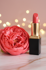 L'Paige lipstick tube in the color Mango  beside a large coral peony flower on a light marble surface, with warm bokeh lights in the background.