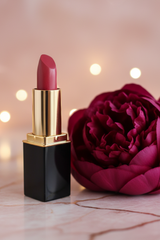A lipstick tube by L'Paige in the color #58 Raspberry  beside a large deep pink flower on a light marble surface with warm blurred lights in the background.