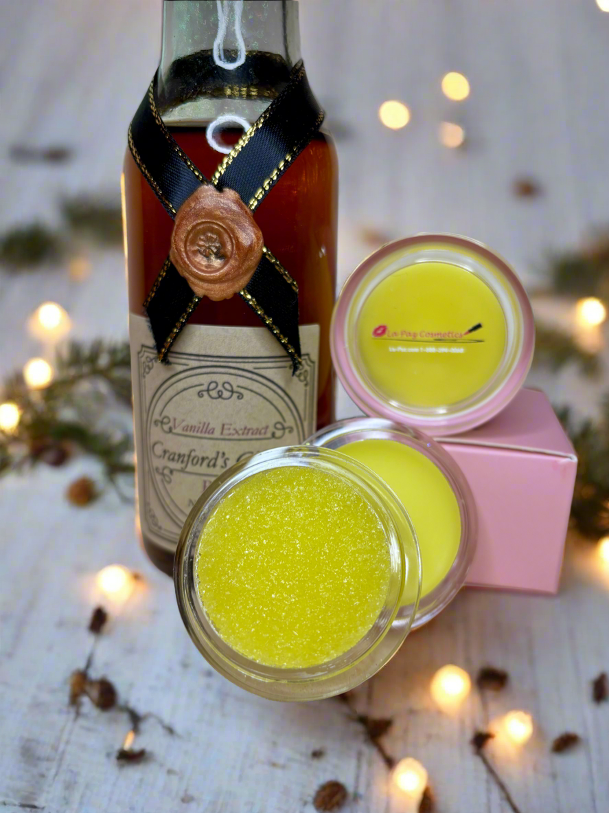 Two jars of yellow vanilla sugar lip scrub with a bottle of vanilla extract on a wooden surface with blurred lights in the background.