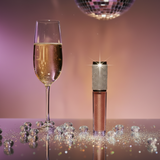 Champagne flute with sparkling Champagne Rose matte lipstick and disco ball on a reflective surface with a purple gradient background.