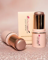 La Paz Cosmetics Adorned Glow 05 Shimmer Highlighter Stick — two gold and soft pink sticks open beside the La Paz Cosmetics box on a pink glitter surface with soft warm bokeh in the background, showing the soft mauve pearl shimmer formula