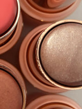 Close-up of La Paz Cosmetics Adorned Glow 05 Shimmer Highlighter Stick showing the soft mauve pearl shimmer formula in a rose gold case, centered among other shades in the collection including blush and bronzer sticks