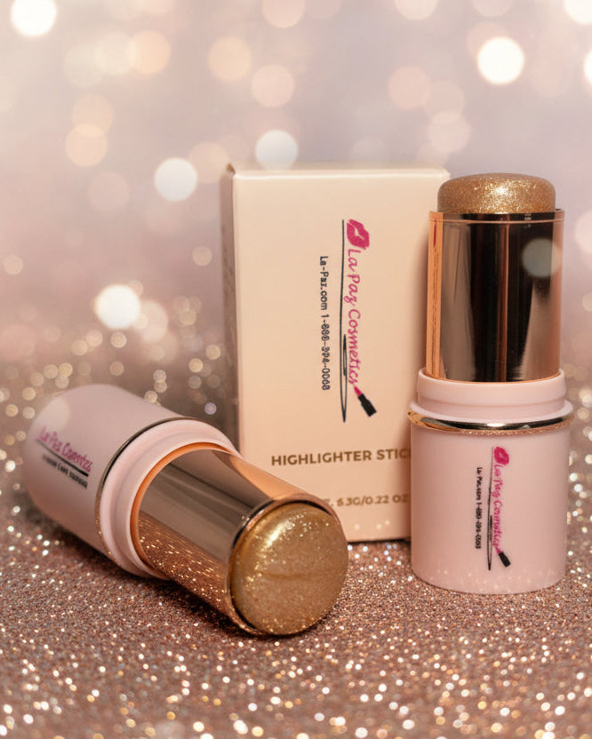 La Paz Cosmetics Angelic Glow 12 Glitter Highlighter Stick — two gold and soft pink sticks open and  beside the La Paz Cosmetics box on a sparkling gold glitter surface with warm champagne bokeh fairy lights in the background