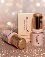 La Paz Cosmetics Angelic Glow 12 Glitter Highlighter Stick — two gold and soft pink sticks open and  beside the La Paz Cosmetics box on a sparkling gold glitter surface with warm champagne bokeh fairy lights in the background