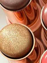 Extreme close-up of La Paz Cosmetics Angelic Glow 12 Glitter Highlighter Stick showing the chunky champagne gold glitter texture of the formula in a rose gold case, surrounded by other shades in the collection