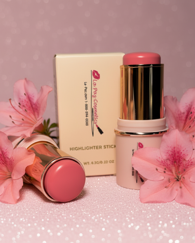 La Paz Cosmetics Cream Blush Stick in Azalea 03 — two gold and soft pink sticks opened beside the La Paz Cosmetics box, surrounded by fresh pink azalea flowers on a pink glitter background"