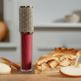 Lip gloss with rhinestone top on a wooden surface next to a slice of apple pie and an apple.