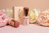 La Paz Cosmetics Cream Blush Stick in Buttercup 04 — two gold and cream sticks open and closed beside the La Paz Cosmetics box, surrounded by white and blush peony flowers on a pink glitter background