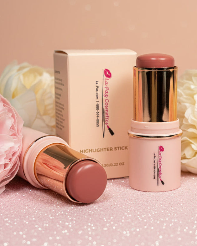 La Paz Cosmetics Cream Blush Stick in Buttercup 04 — two gold and cream sticks open and closed beside the La Paz Cosmetics box, surrounded by white and blush peony flowers on a warm beige background