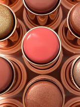 Overhead close-up of La Paz Cosmetics Cream Blush and Highlighter Sticks in gold cases, with Azalea 03 — a vibrant rosy pink — centered among the full collection of shades including bronzers and highlighters