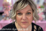 Woman with styled hair and makeup in a blurred indoor setting with text 'Watermelon clean clear lip oil'.