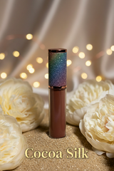 La Paz Cosmetics Cocoa Silk Lux Lip Gloss S21 — rich cocoa bronze gloss with holographic rainbow glitter cap, styled with white peonies on a gold glitter surface with warm bokeh fairy lights and ivory silk draping