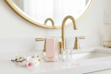 Bathroom vanity with gold fixtures, a pink bottle, and small flowers.
