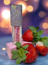 Clear Strawberry lip oil with rhinestone top on a pink box, surrounded by strawberries against a blurred background.