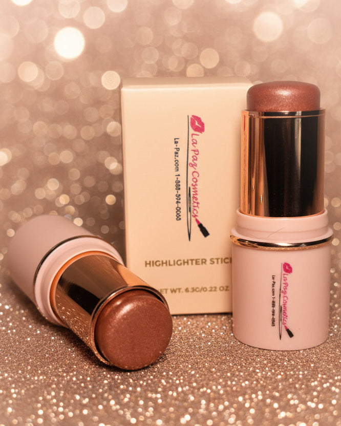 La Paz Cosmetics Sun Kissed Glow 8 Shimmer Bronzer Stick — two gold and cream sticks open and closed beside the La Paz Cosmetics box on a sparkling gold glitter surface with warm champagne bokeh in the background, showing the warm bronze shimmer formula