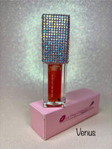A tube of Venus Magic glowing lip oil by La Paz Cosmetics, displayed in a clear container with a sparkling, multicolored lid, set against a pink backdrop.