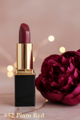 A L'Paige lipstick in color Plum Red with black tube, standing on a marble surface next to a large dark pink peony flower.