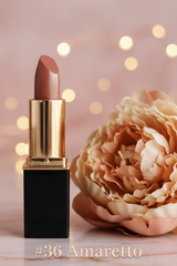 A lube of #36 Amaretto lipstick with a black and gold tube stands on a light marble surface beside a large peach flower