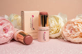 La Paz Cosmetics Cream Blush Stick in Buttercup 04 — two gold and cream sticks open and closed beside the La Paz Cosmetics box, surrounded by white and blush peony flowers on a pink glitter background