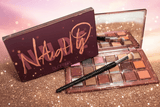 Naughty Nude Eyeshadow Palette open next to its burgundy box on a gold glitter surface with a rhinestone bling eyeshadow brush
