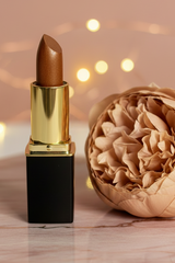 #05 Frosted Copper lipstick with a gold and black tube stands on a pink marble surface next to a dried beige flower, with warm bokeh string lights in the background.