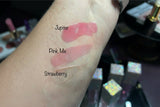Lip oil swatches on a wrist labeled 'Jupiter', 'Pink Me', and 'Strawberry' with makeup products in the background.