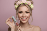 Model holding La Paz Crystal Frosting Lip Gloss Honeyed Rosebud D12 with sheer pink shimmer lips and floral icing crown