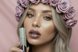 Model holding La Paz Crystal Frosting Lip Gloss in Sugarplum Fairy with sheer plum shimmer lips on pink background