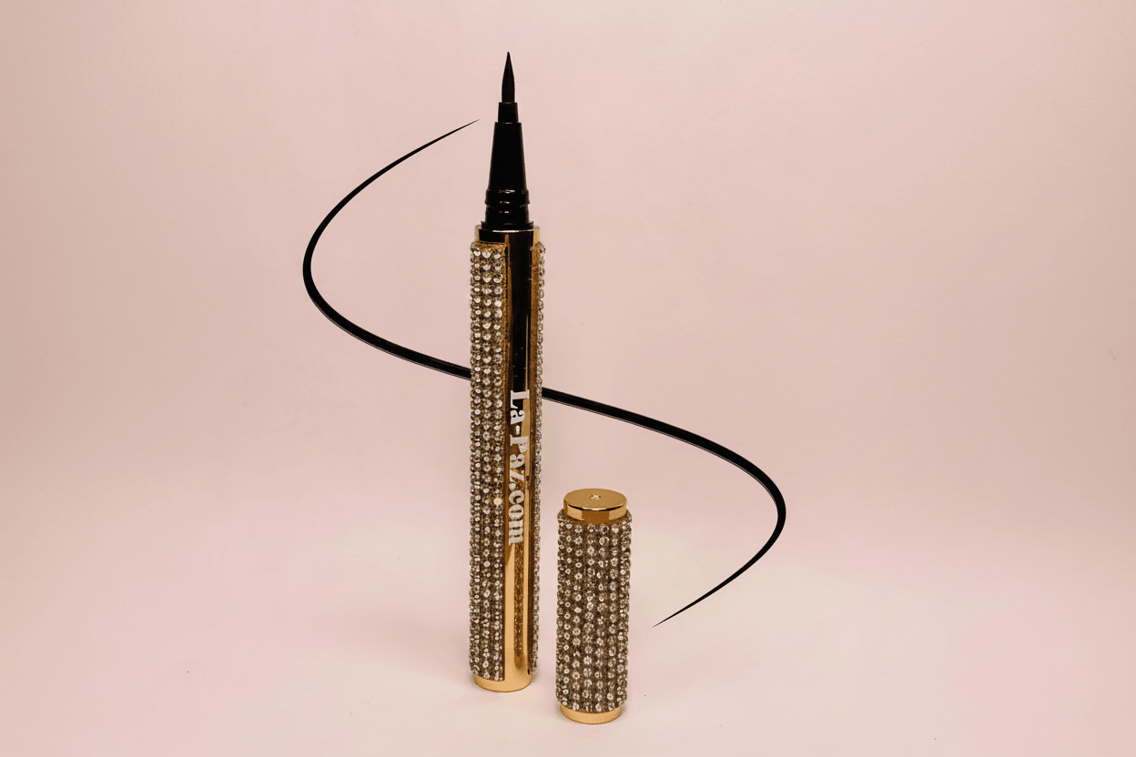 La Paz Cosmetics Luxe Felt Tip Liquid Eyeliner in Black — rhinestone and gold pen open with ultra-fine felt tip displayed beside a dramatic black swoosh line on a blush pink background