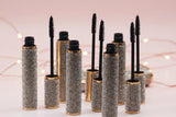 Five La Paz Cosmetics Luxe Lengthening Mascara tubes with rhinestone-encrusted silver and gold cases open with wands displayed on a reflective surface with warm fairy lights on a blush pink background