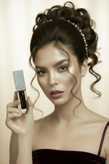 A woman with styled brown hair and a rhinestone headband holds a rhinestone-embellished Black Lip Oil tube against a plain gray studio background.