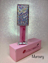 A tube of Mercury Magic glowing lip oil displayed in front of its pink packaging box, with the brand name 'La Paz Cosmetics' visible on the box. The background is glittery.