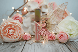 La Paz Pixie Dust Lux Lip Gloss — warm pink gloss with rainbow cap, fairy figurine & pink roses on wood with fairy lights