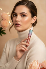 Woman in cream cashmere sweater holding La Paz Lux Lip Gloss in Cashmere Peach with iridescent cap on soft beige floral bokeh