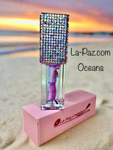 Oceans lip oil with a glittery rhinestone cap sitting on a beach at sunset.