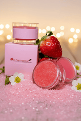 La Paz Cosmetics Strawberry Lip Sugar Scrub and Balm set — open scrub jar showing pink sugar crystals and lip balm jar beside a pink La Paz gift box, fresh strawberry, and white strawberry blossoms nestled in a bed of pink sugar crystals with warm bokeh fairy lights