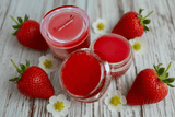 A container of La Paz Cosmetics Strawberry Lip Sugar Scrub and Balm  with another container open showing the two different sides on a rustic white wood surface surrounded by fresh red strawberries and white strawberry blossoms