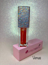 A tube of Venus Magic glowing lip oil by La Paz Cosmetics, displayed in a clear container with a sparkling, multicolored lid, set against a pink backdrop.